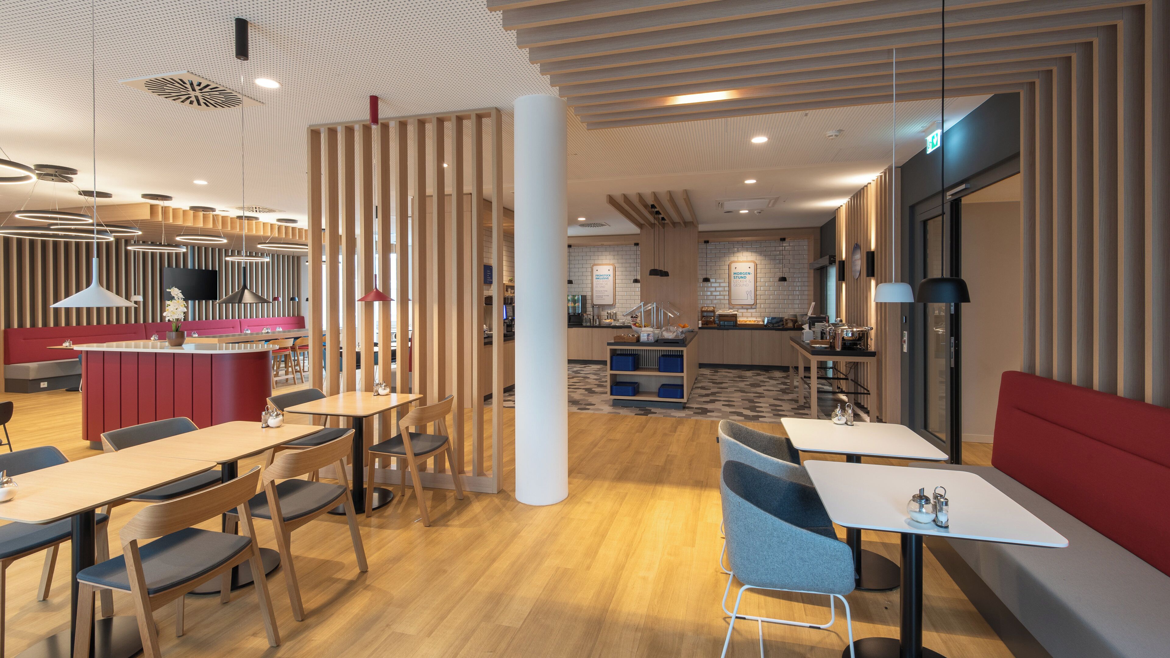 A modern restaurant with wooden floors, red seating, and a kitchen area in the background.