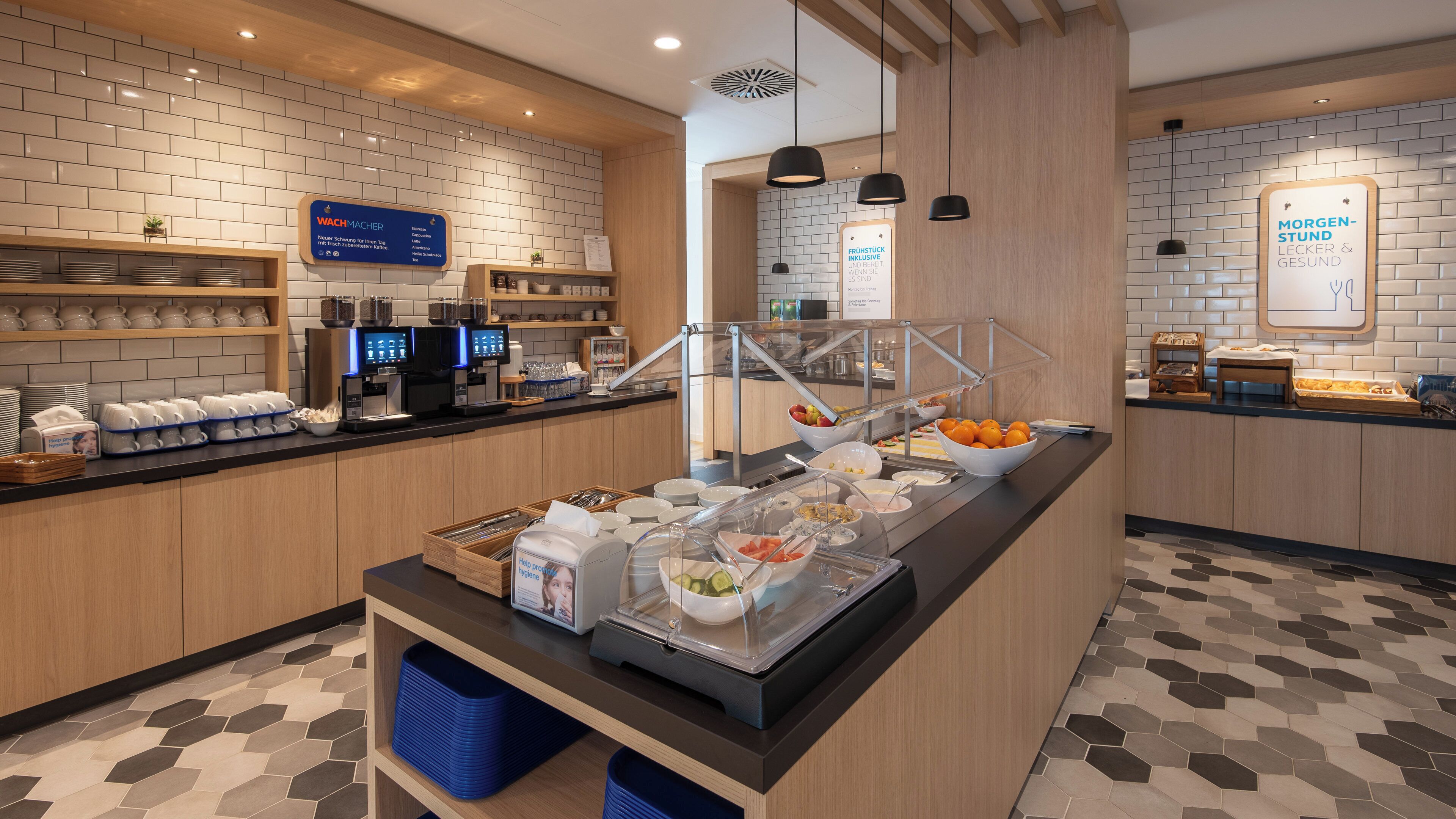 A breakfast buffet area with coffee machines, fruit bowls, and a variety of breakfast items.