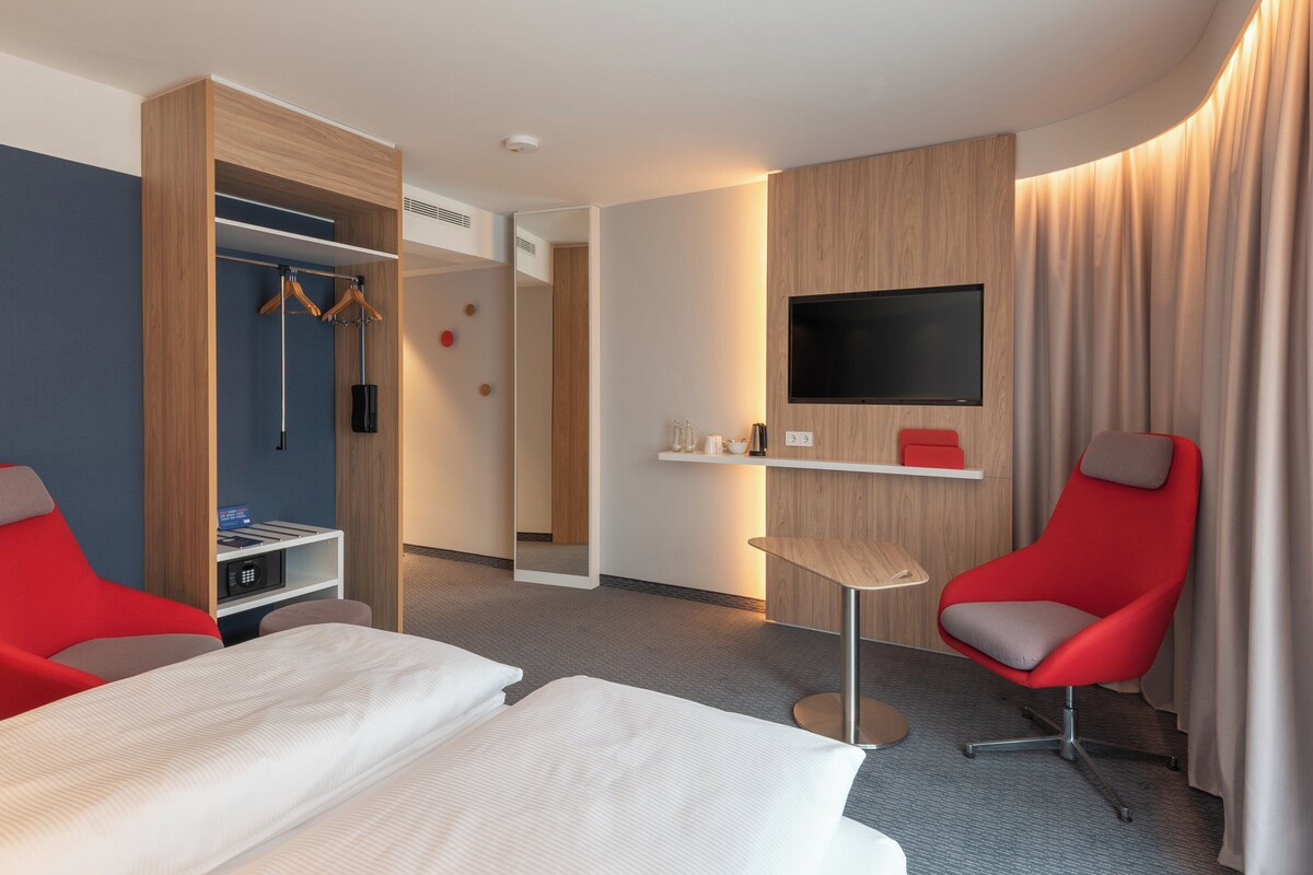 Une chambre d’hôtel avec un lit, deux chaises rouges, une télévision, un miroir, une armoire et une petite table.