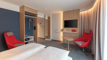 Ein Hotelzimmer mit einem Bett, zwei roten Sessel, einem Fernseher, einem Spiegel, einem Kleiderschrank und einem kleinen Tisch.