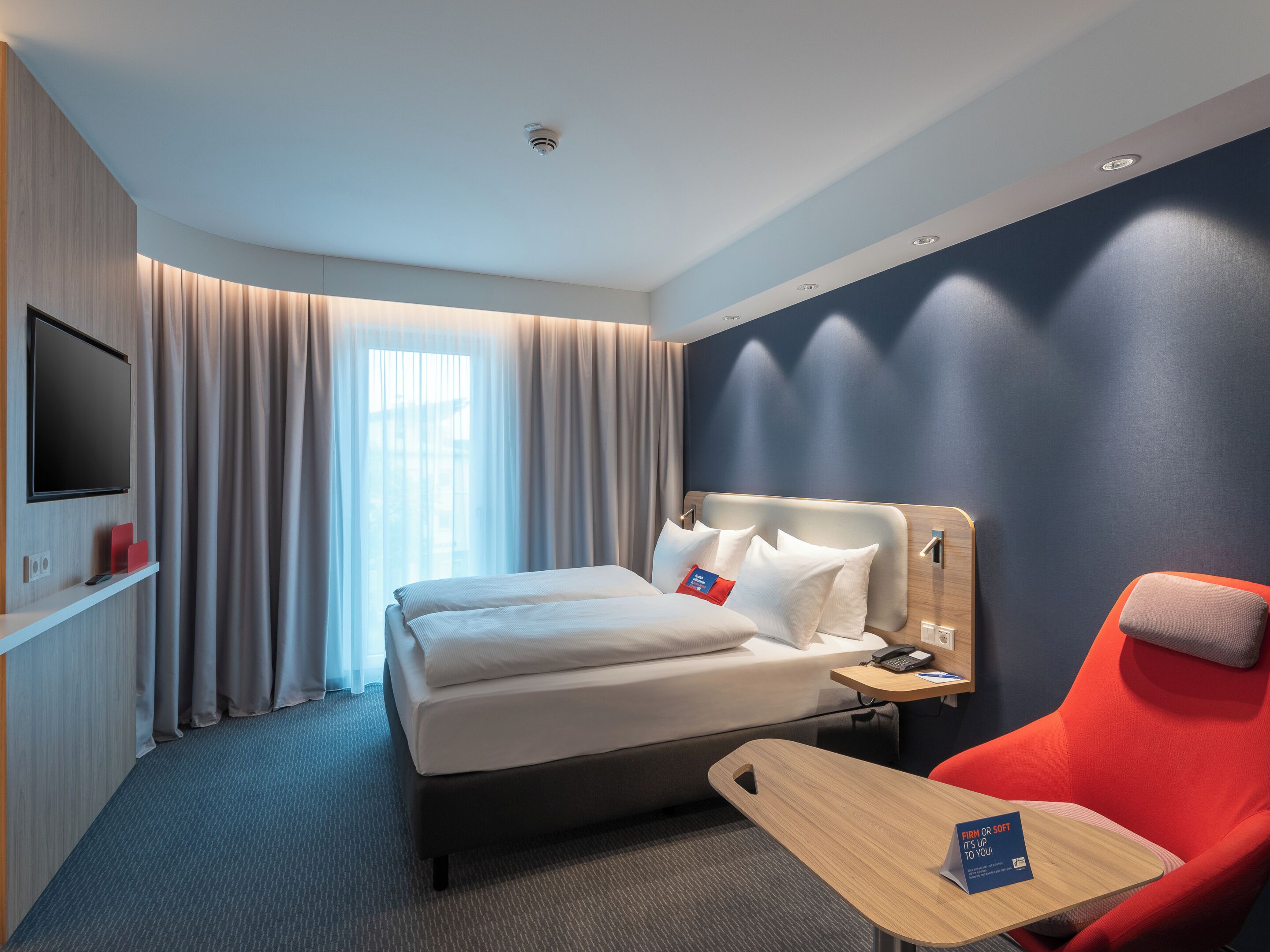 Une chambre d’hôtel avec un lit, un bureau, une chaise rouge, une télévision et des rideaux.