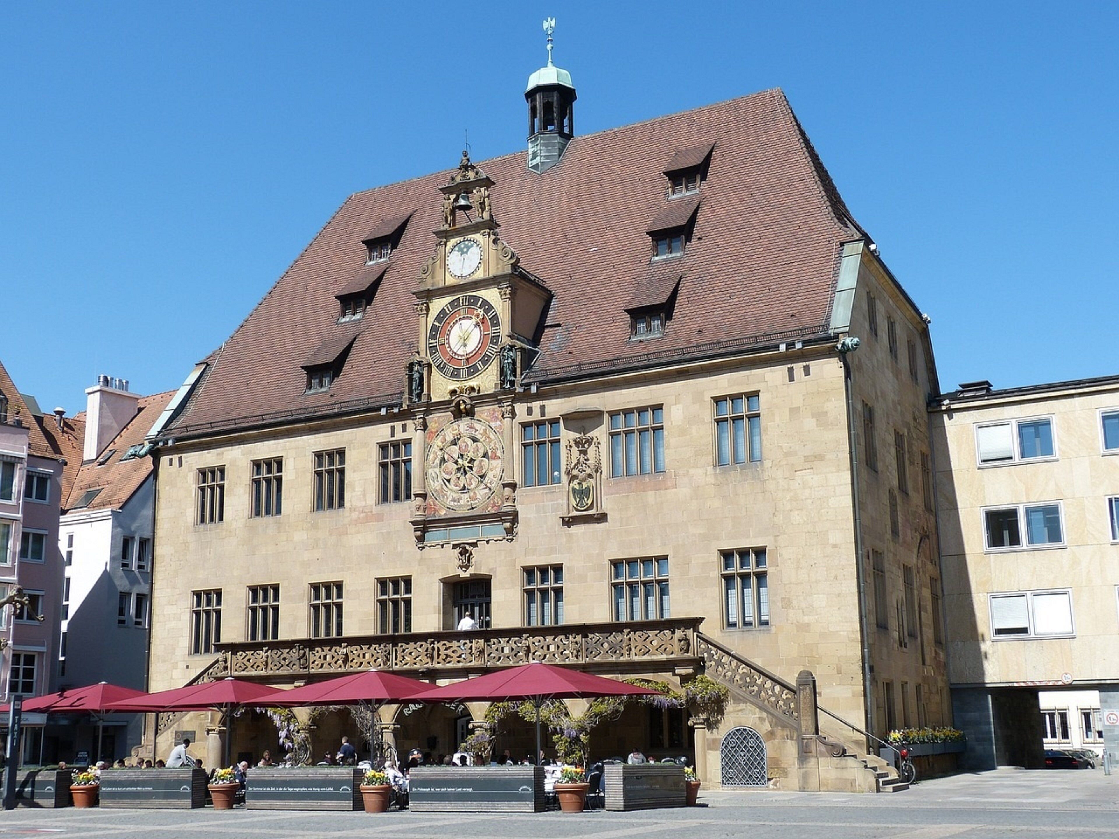 A historic building with a prominent clock tower, large windows, and a covered outdoor seating area with red umbrellas.