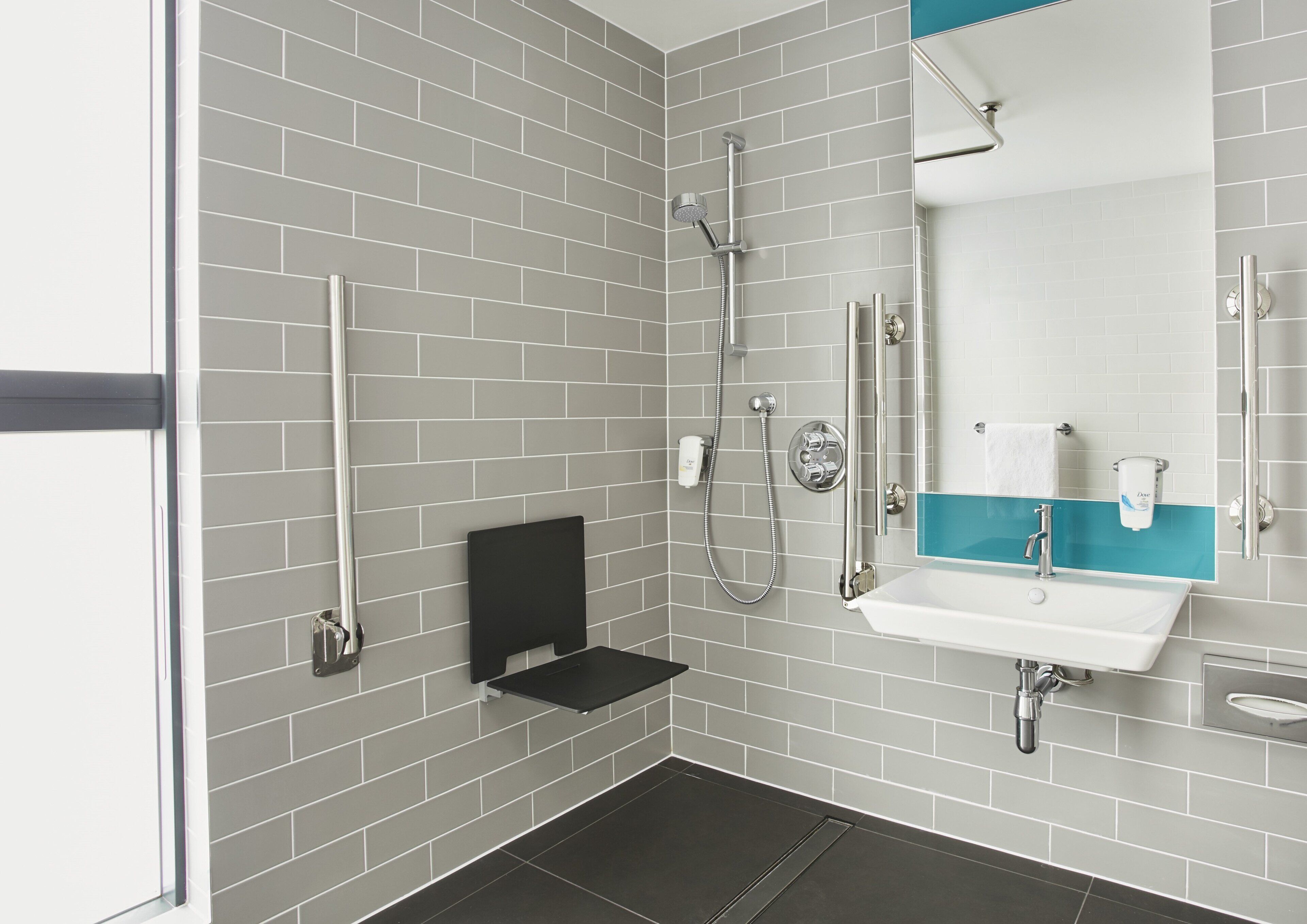 Une salle de bain moderne avec des murs recouverts de carreaux gris, une étagère noire, un lavabo blanc agrémenté d’une touche de bleu et une douchette à main.