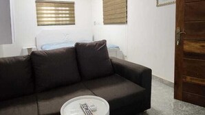 A living room with a dark brown sofa, a small round coffee table, a ceiling fan, and a door on the right. - 696 Arena Aflao (Aflao)