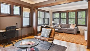 A spacious living room with a sofa, armchair, and coffee table. There are large windows with blinds, a desk with a chair, and a lamp. - Just A Short Stroll to Lake Harriet & Restaurants! (Minneapolis)
