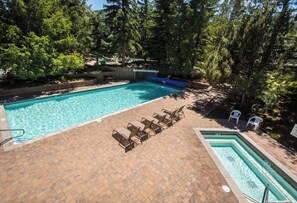 A rectangular swimming pool with a brick deck, surrounded by trees. There are lounge chairs and a small hot tub.