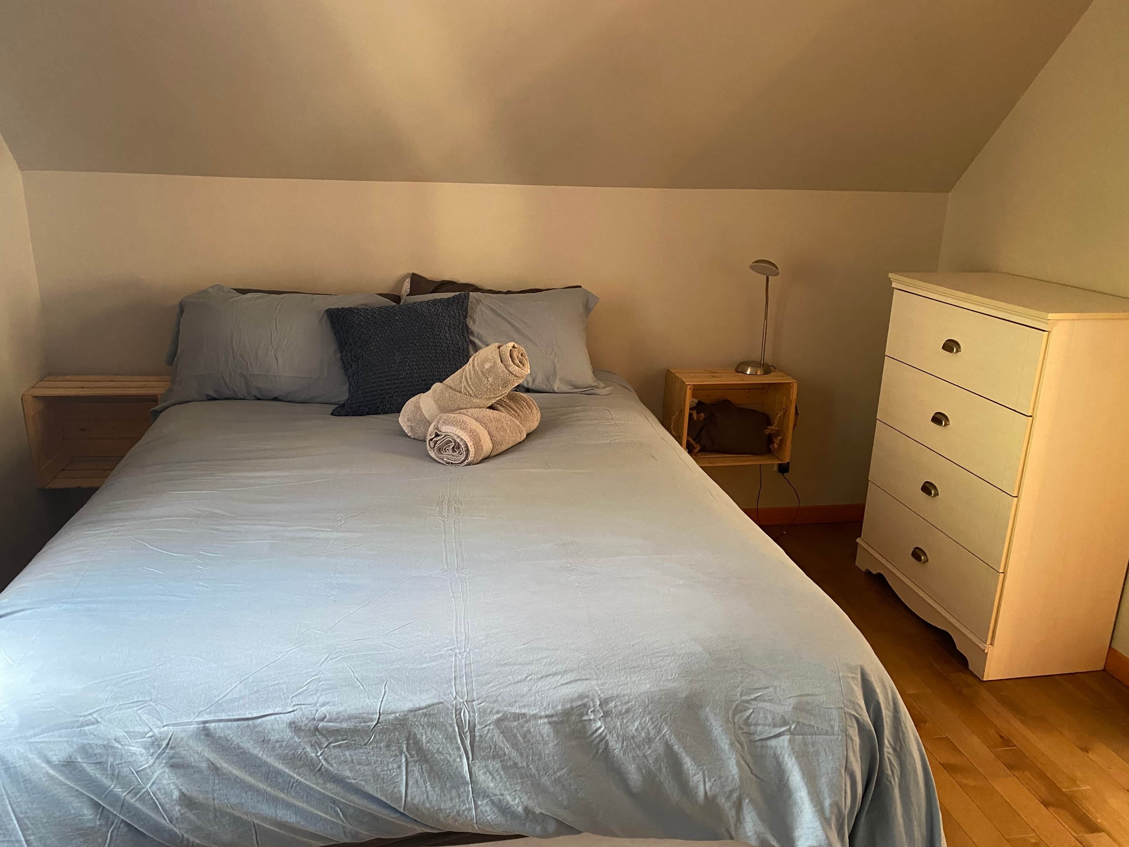 Une chambre à coucher avec un lit, des oreillers, une table de chevet en bois, une lampe et une commode.