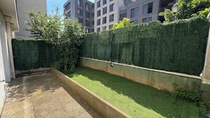 Ein schmaler Innenhof mit Kunstgrasanlage, einem Betonweg und einer hohen Hecke.
