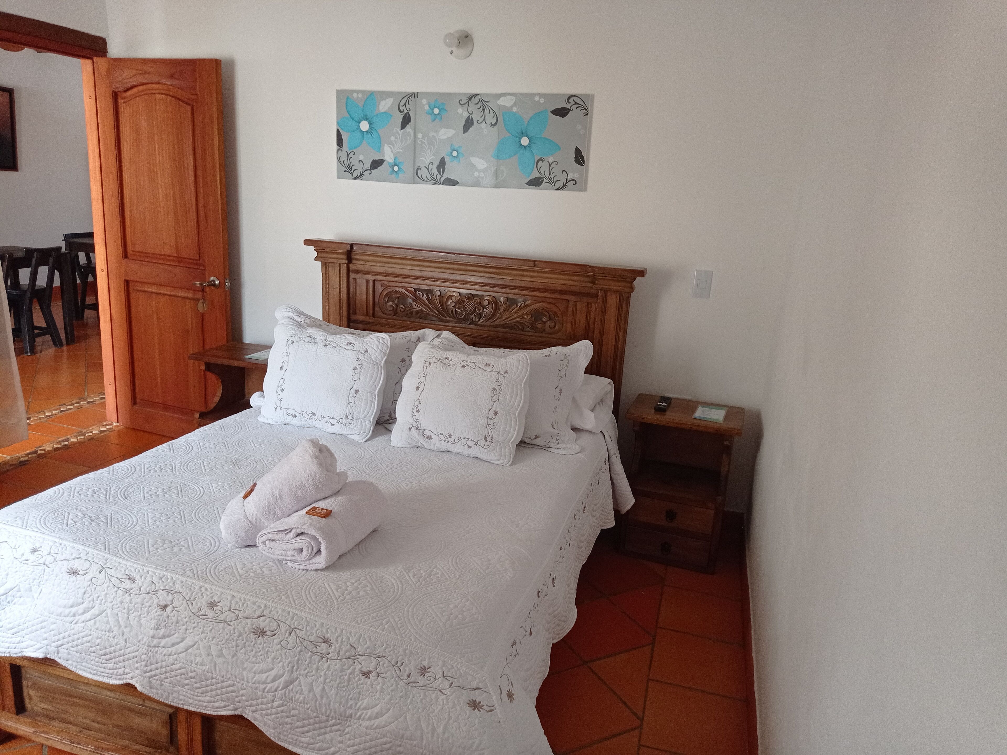 Una cama bien hecha con colcha y almohadas blancas, un cabecero de madera, una mesita de noche con control remoto y una obra de arte en la pared sobre la cama.