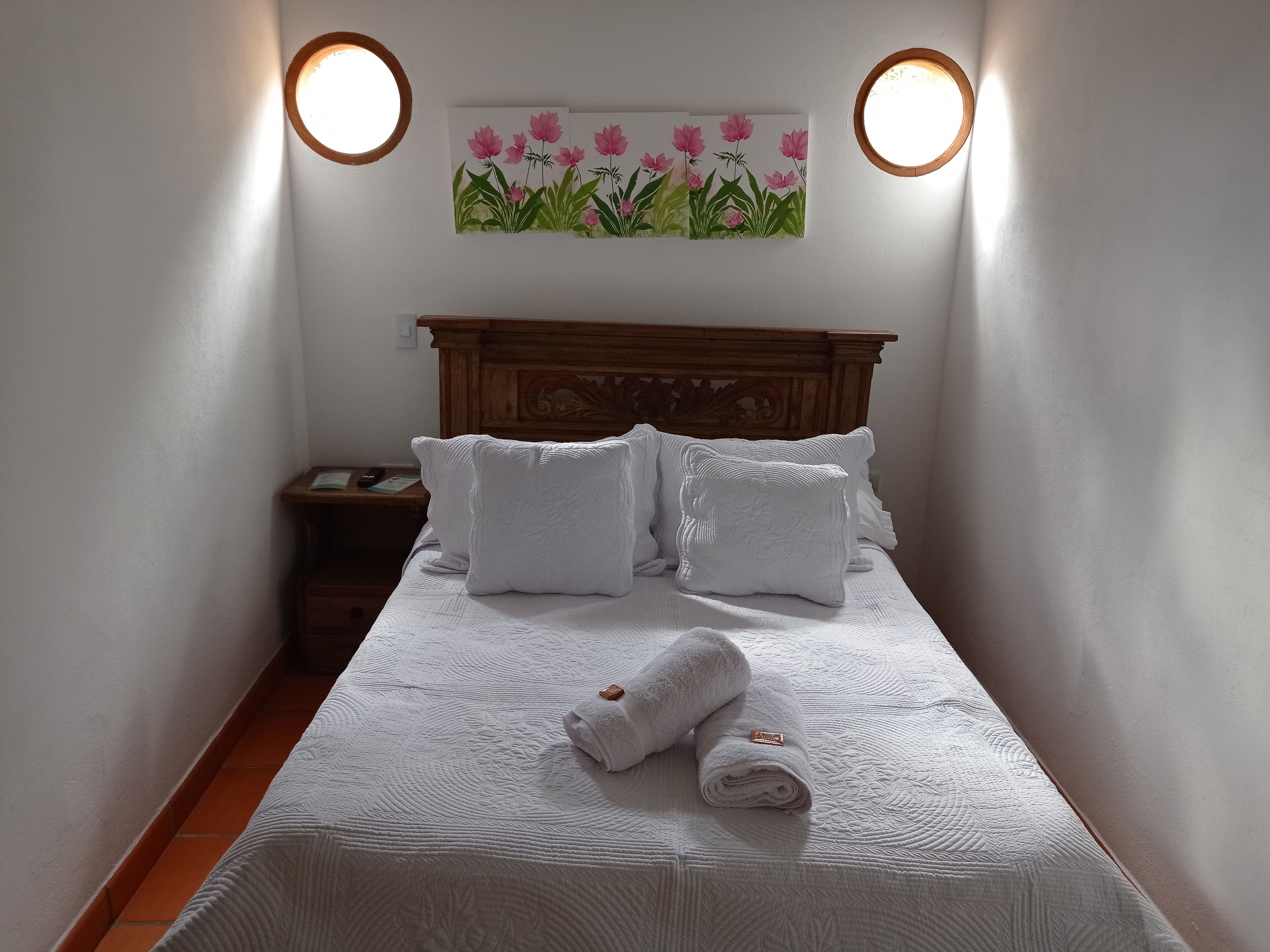 A neatly made bed with white bedding and pillows, a wooden headboard, and a painting of pink flowers on the wall above.