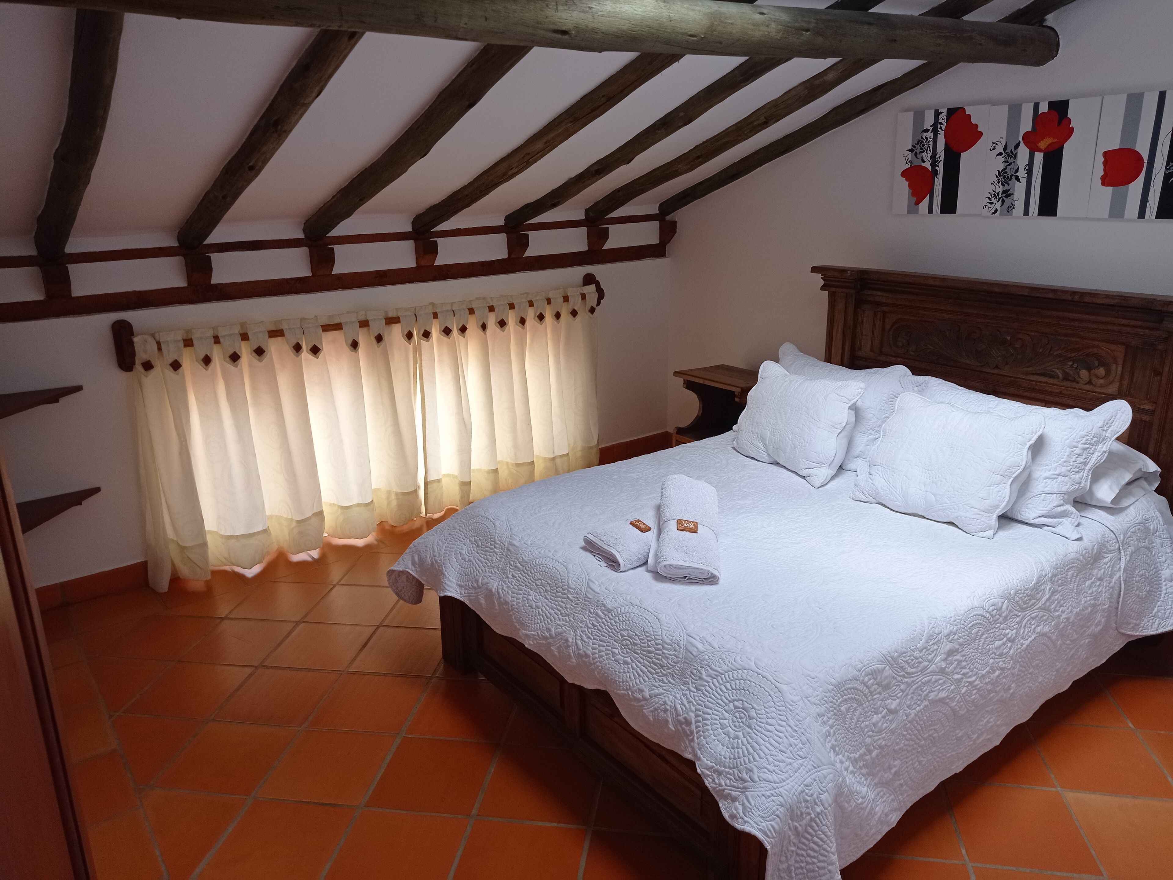 Un dormitorio con cama de madera, ropa de cama blanca, cabecera de madera, mesita de noche y una ventana con cortinas.