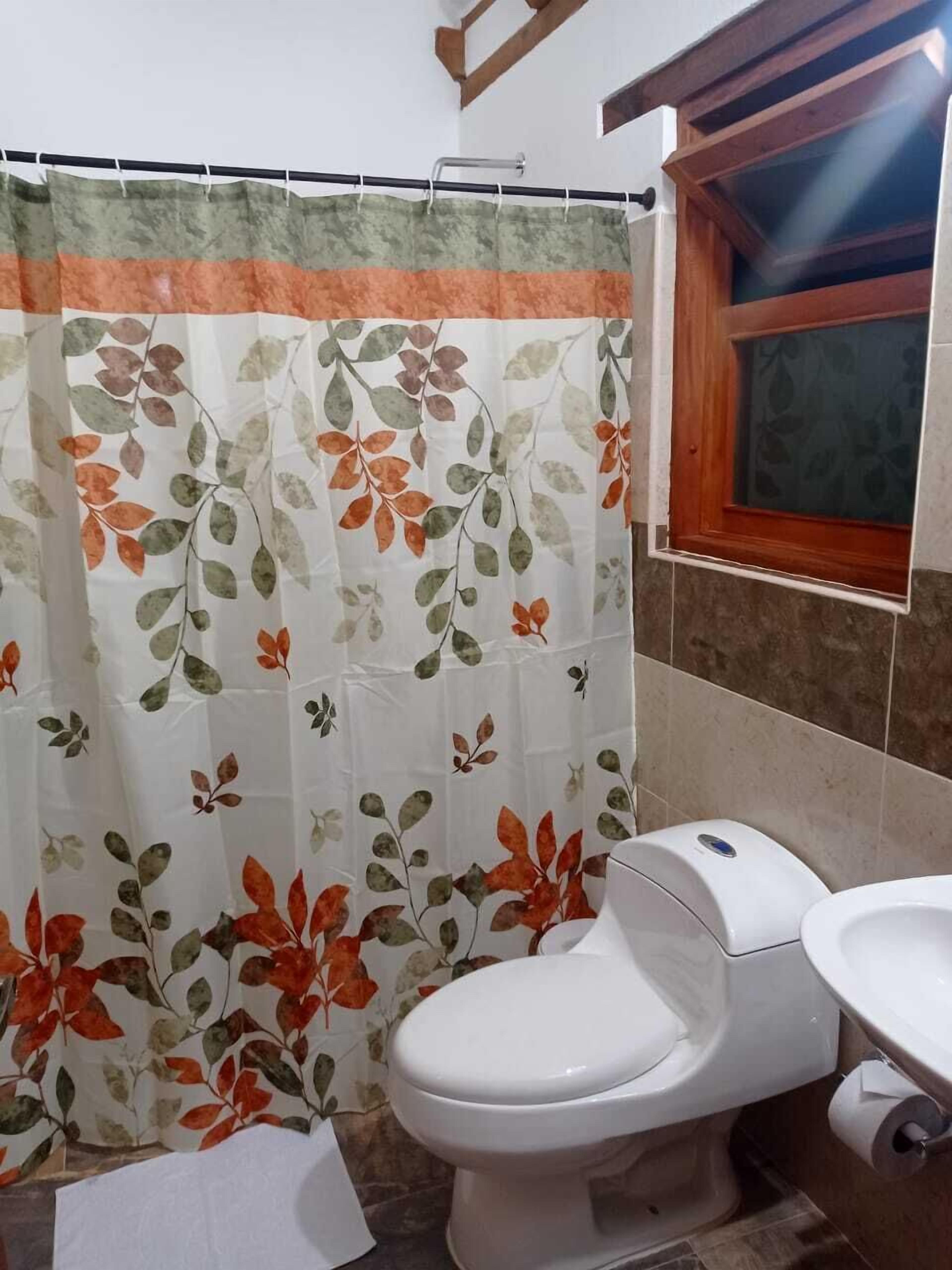 A bathroom with a toilet, a shower curtain with a leaf pattern, and a window with wooden frames.