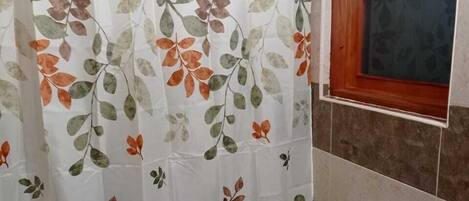 A bathroom with a toilet, a shower curtain with a leaf pattern, and a window with wooden frames.