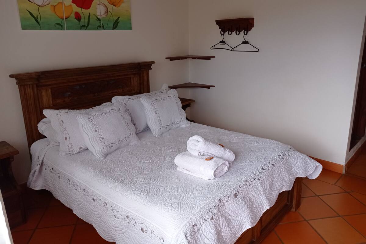 Un lit bien fait, recouvert d’une couette blanche, avec deux oreillers et une serviette enroulée. On y voit une tête de lit en bois, un porte-manteau fixé au mur et un tableau de fleurs au-dessus du lit.
