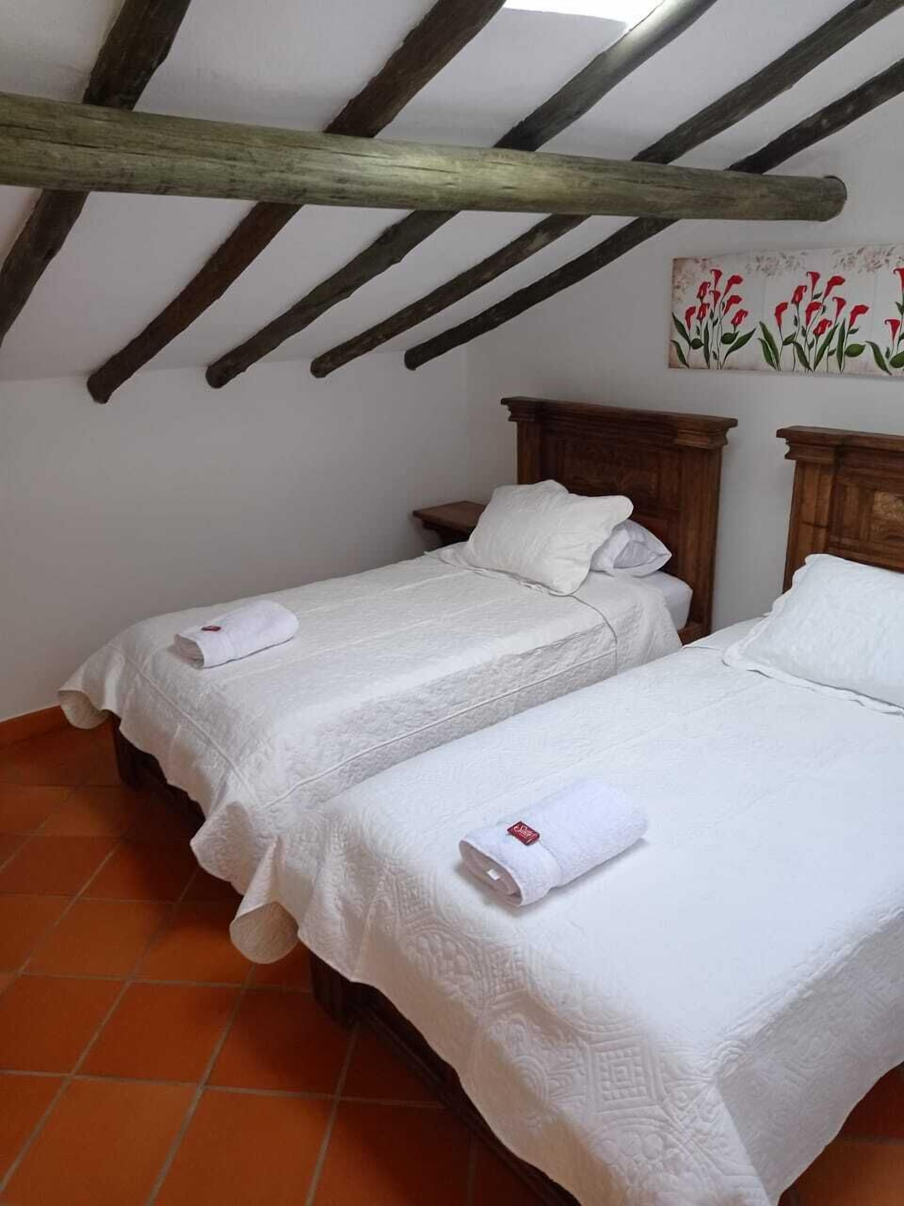 A room with two beds, each with a white bedspread and a rolled towel. The room has a sloped ceiling with wooden beams and a tiled floor.