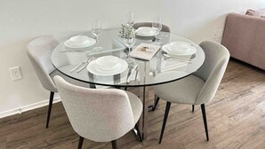Mesa de vidro redonda com quatro cadeiras cinzas estofadas, pratos brancos, talheres e copos de vinho dispostos para quatro pessoas.