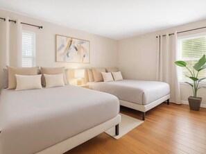 Ein Schlafzimmer mit zwei Betten, einem Nachttisch, einer Lampe, einem Fenster mit Vorhängen und einer Zimmerpflanze.