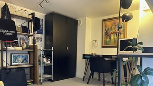 Ein Zimmer mit Schreibtisch, Stuhl, Regalen mit verschiedenen Gegenständen, einem schwarzen Kleiderschrank und einem Bild an der Wand.