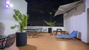 A rooftop terrace with a potted plant, outdoor furniture, and a view of the city at night.