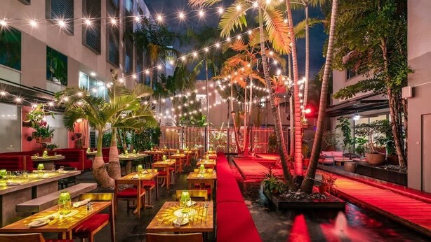 Un área de comedor al aire libre con mesas, sillas, luces colgantes y palmeras.