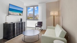 Una habitación de hotel moderna con un ventanal grande, un televisor de pantalla plana, un mueble de almacenamiento negro, un sofá blanco y una mesita de centro redonda.
