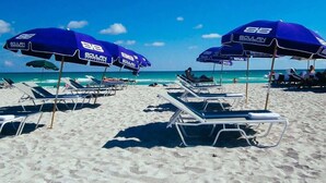 Una playa con filas de tumbonas y sombrillas azules, con la marca "BOULAN" a la vista.