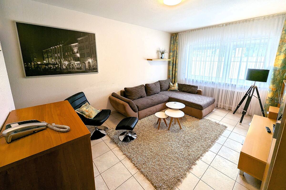 Una sala de estar con un sofá modular, dos mesas redondas pequeñas, un teléfono y un televisor sobre un trípode.