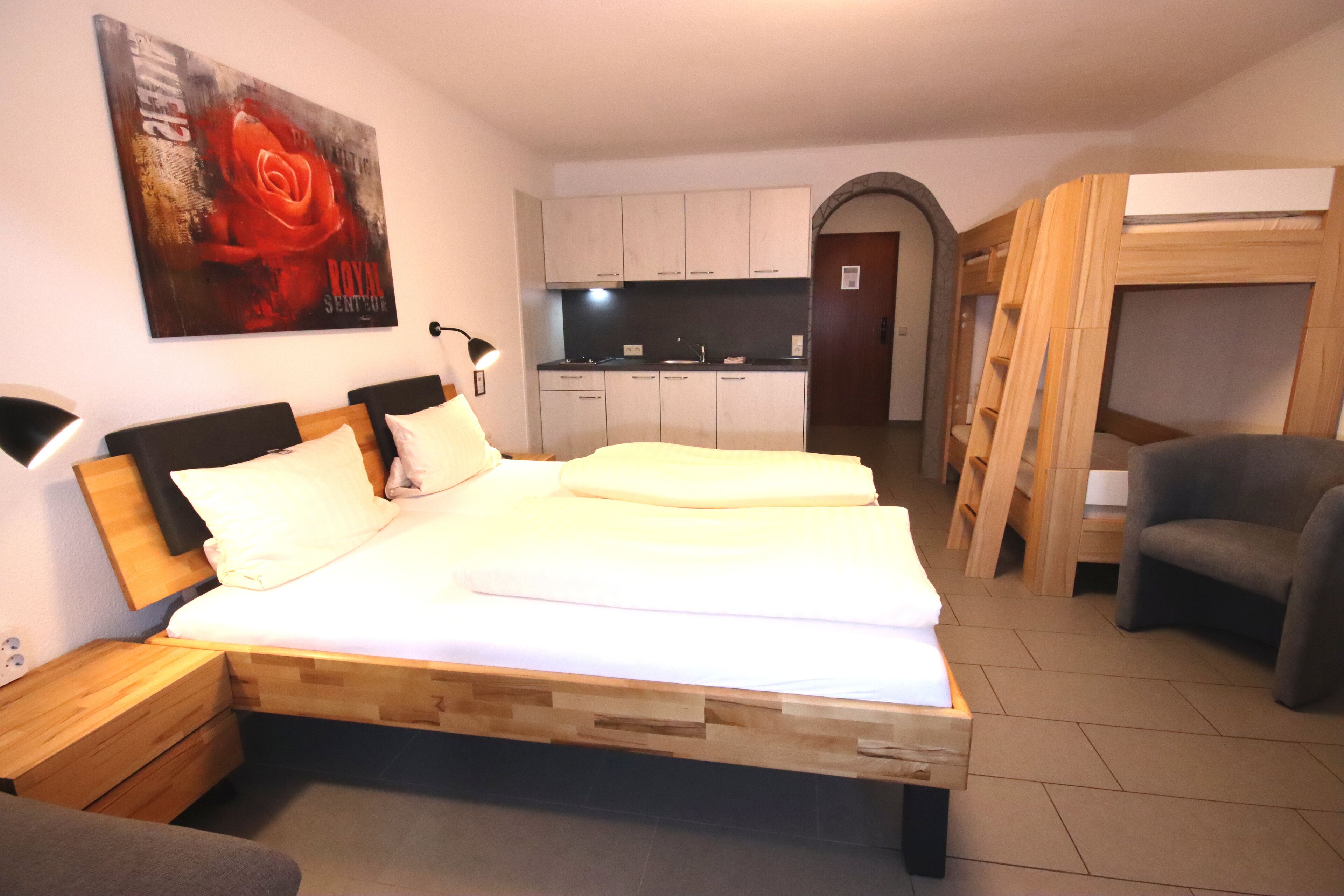 Ein Schlafzimmer mit einem hölzernen Bett, einem Nachttisch, einem Schreibtisch, einem Stuhl und einer kleinen Küche im Hintergrund.