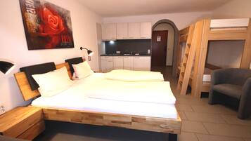 Ein Schlafzimmer mit einem hölzernen Bett, einem Nachttisch, einem Schreibtisch, einem Stuhl und einer kleinen Küche im Hintergrund.