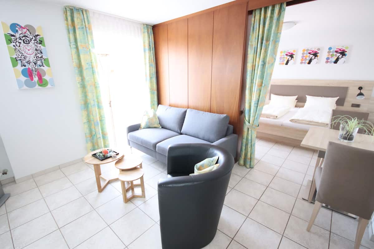 Una sala moderna con un sofá gris, un sillón negro, una mesa de centro de madera y un taburete pequeño de madera. Se observa una cama con ropa blanca de cama al fondo.