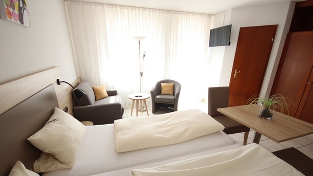 A hotel room with two beds, a sofa, a small table, a chair, a floor lamp, and a TV on the wall. - Appartements Biedermeier (Bad Krozingen)