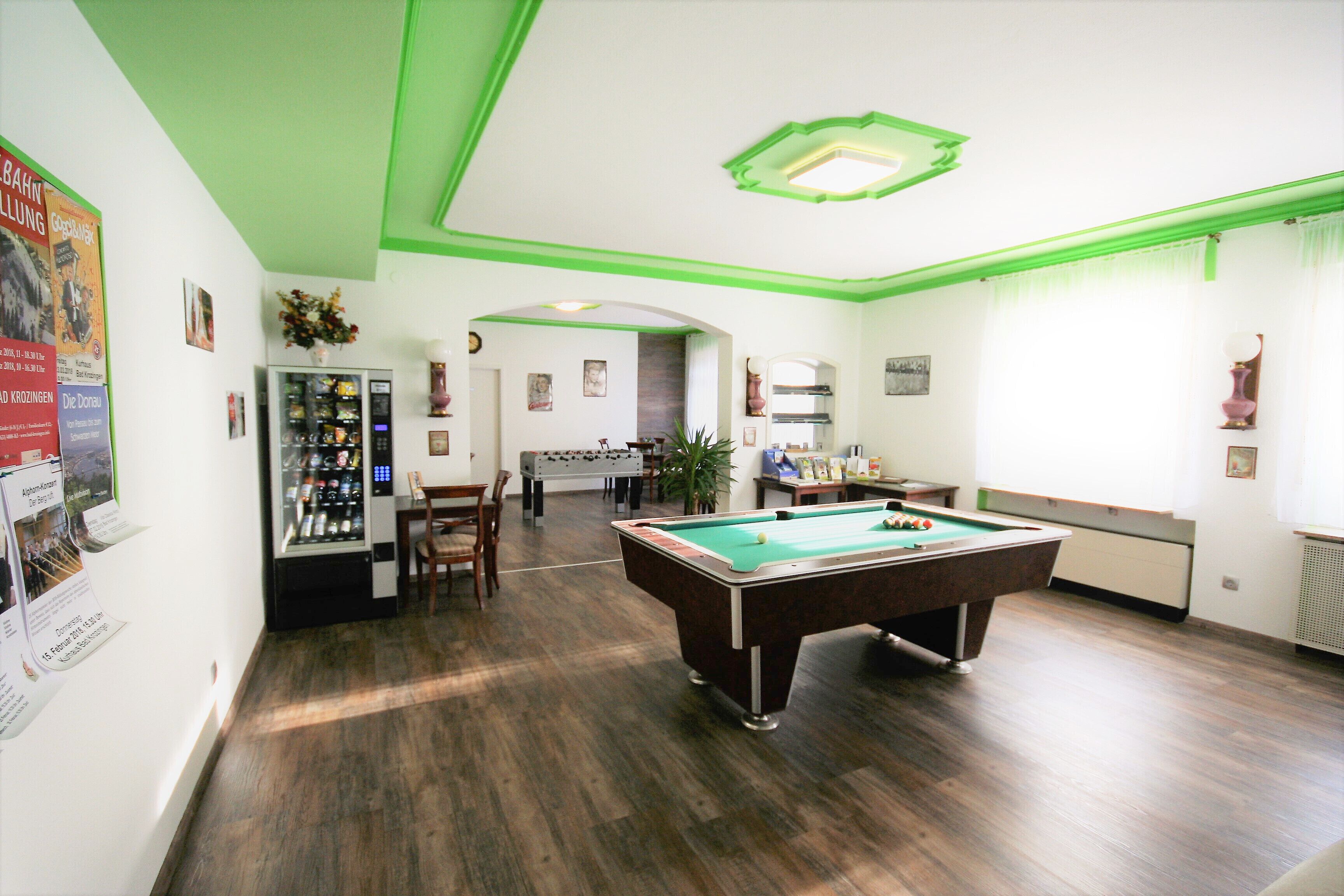 A billiards room with a pool table, a foosball table, and a kitchen area in the background.