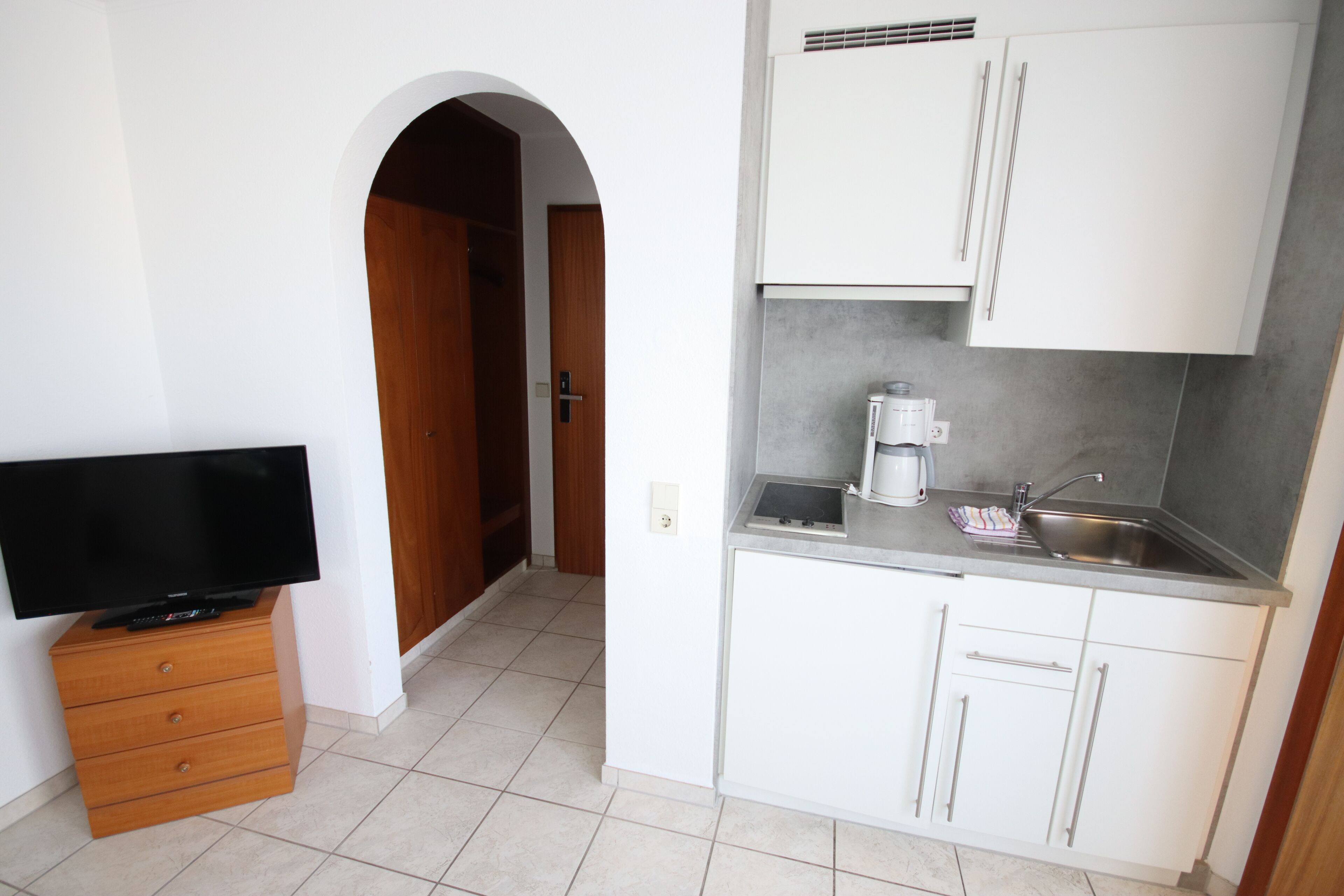 A compact kitchen with white cabinets, a built-in coffee maker, and a flat-screen TV on a wooden cabinet.