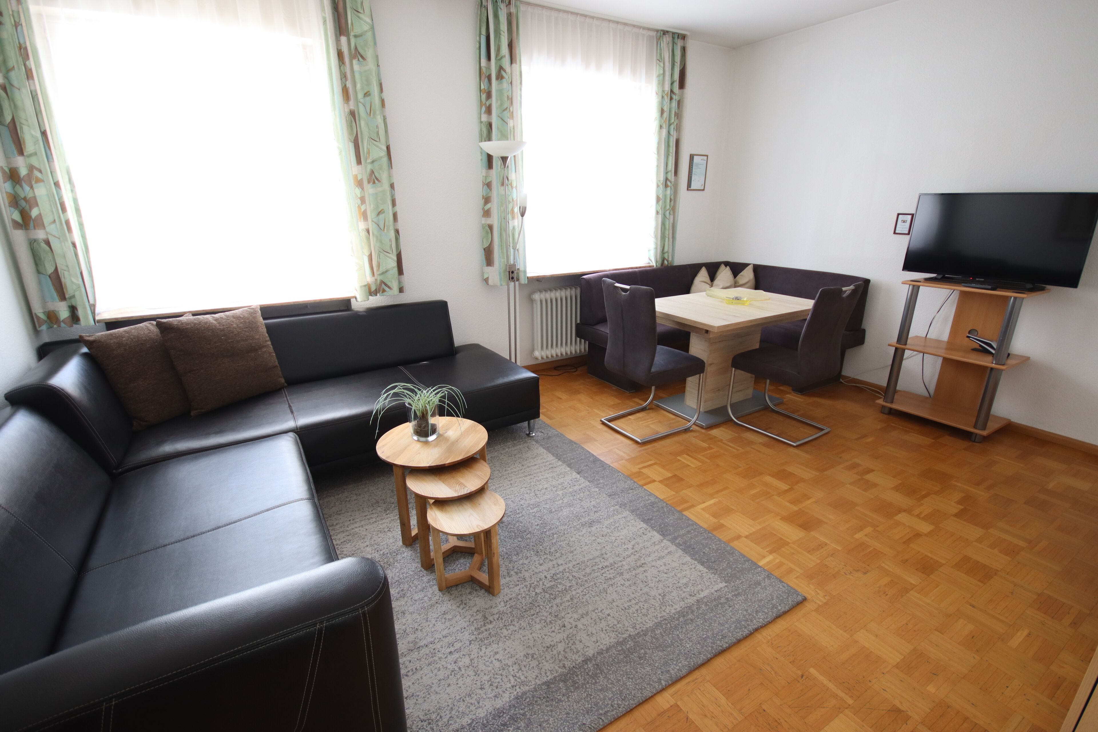 A modern living room with a black leather sofa, a small wooden table with a plant, a dining table with chairs, and a flat-screen TV on a stand.