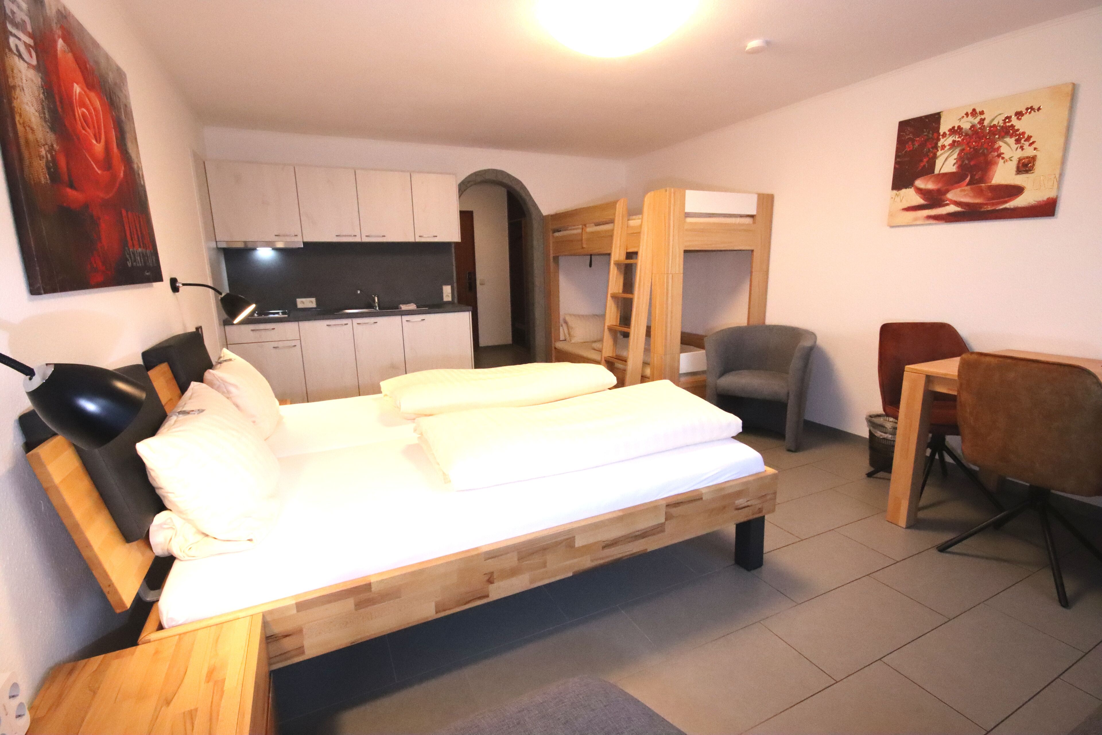 A compact room with a bed, a desk, a chair, a kitchenette, and a painting on the wall.