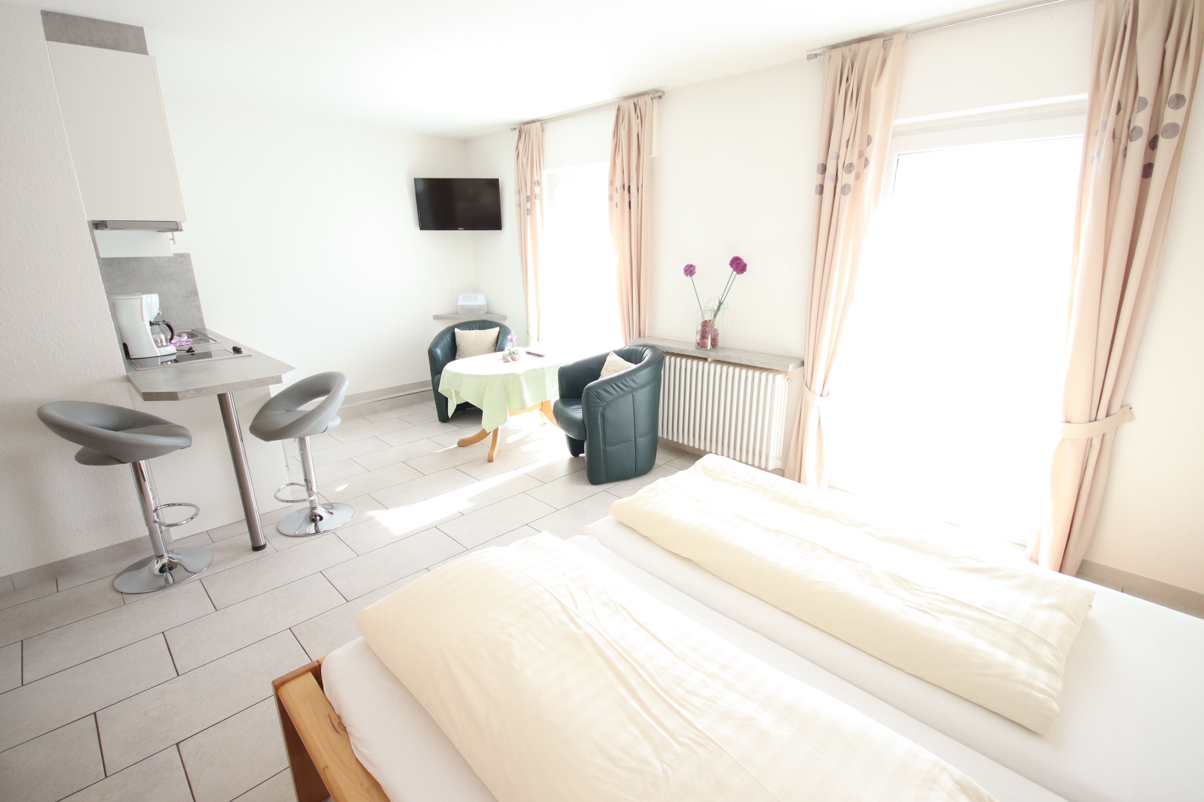 A hotel room with a bed, a desk with a chair, a small table, a radiator, and a TV on the wall.