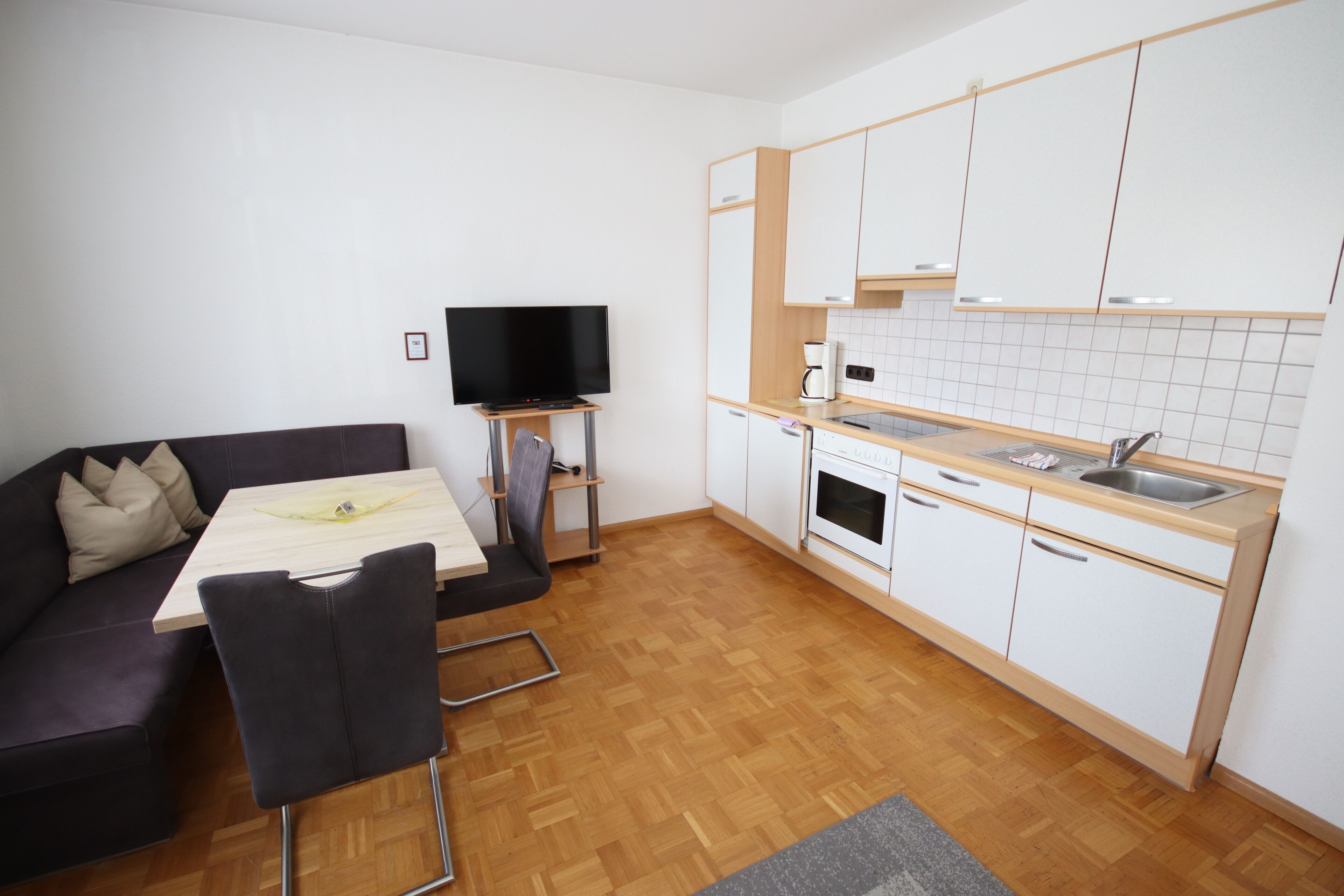 A compact living space with a sofa, a dining table, a TV, a kitchenette, and a wooden floor.