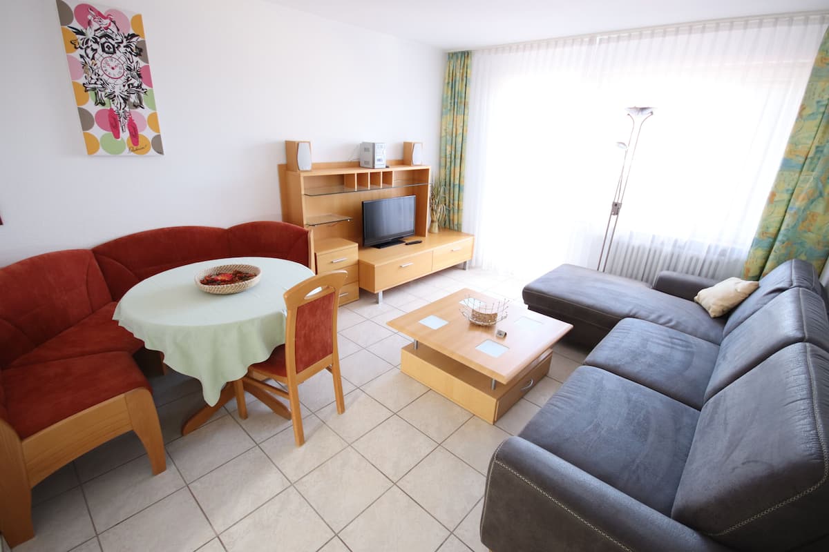 Una sala de estar con un sofá, un televisor sobre un mueble de madera, una mesa de comedor con sillas y una lámpara de pie.