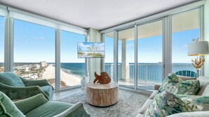 Sala de estar moderna com uma grande janela para uma praia e oceano, uma mesa de centro redonda de madeira, e móveis estofados verdes.