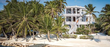Un edificio grande de varios pisos con fachada blanca y ventanas arqueadas, rodeado de palmeras y una playa arenosa.