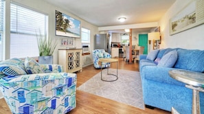 A living room with a blue sofa, patterned ottoman, and a dining area with a green door.