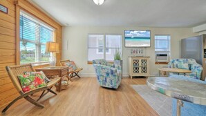 Una sala de estar con pisos de madera, muebles de mimbre, un sofá estampado, un televisor y un mueble aparador.