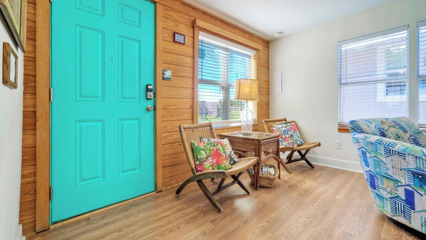 A room with a turquoise door, wooden walls, a wicker chair with a colorful cushion, a wicker table, and a patterned sofa.