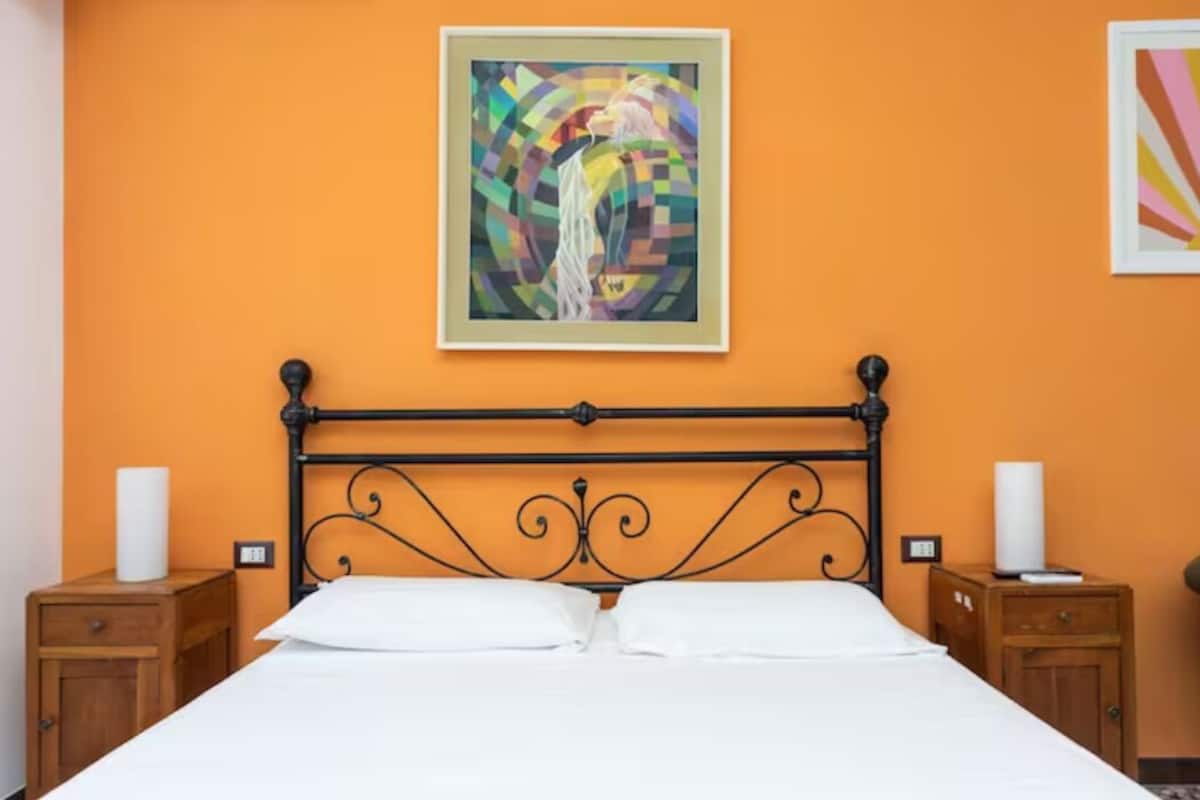 A neatly made bed with a black metal headboard, two wooden nightstands with lamps, and a colorful abstract painting on the orange wall above the bed.