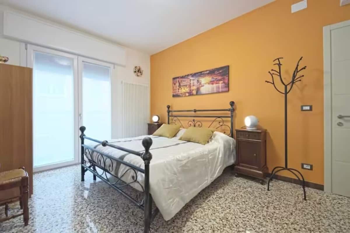 A bedroom with a bed, a nightstand, a chair, a coat rack, and a door.