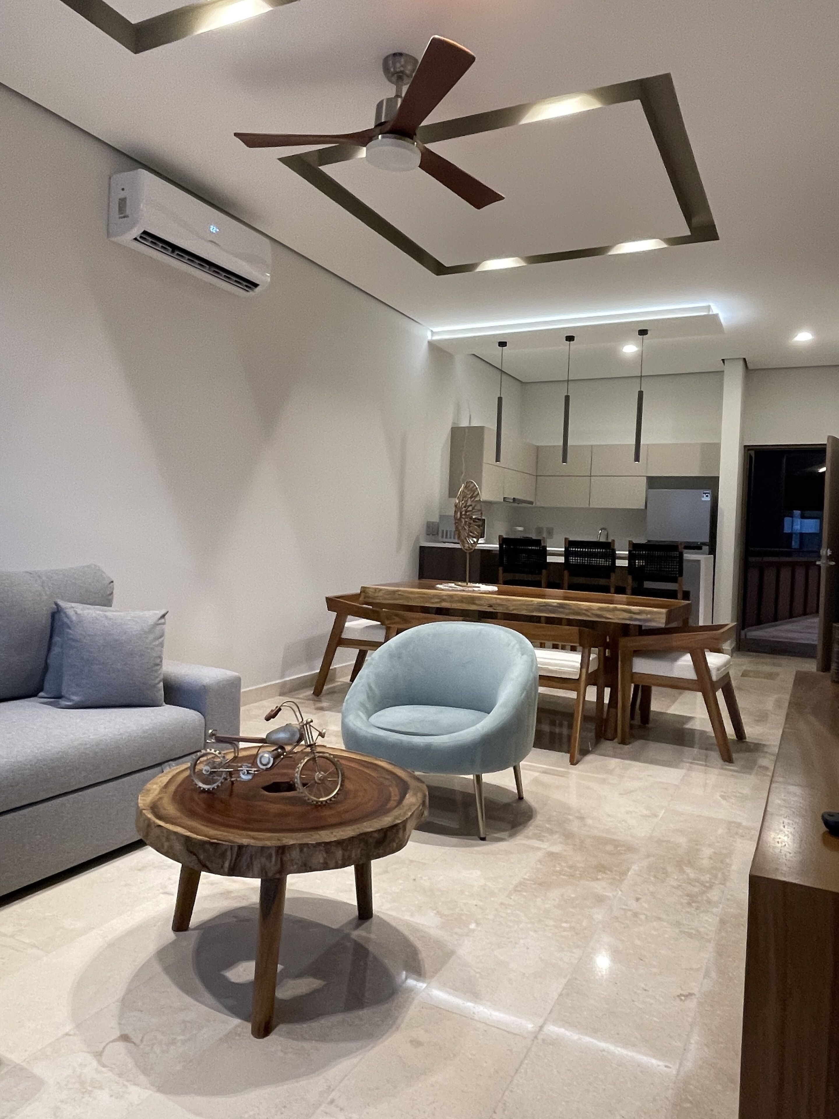 A modern living room with a sofa, a dining table, and a light blue chair. There is a ceiling fan and an air conditioning unit.