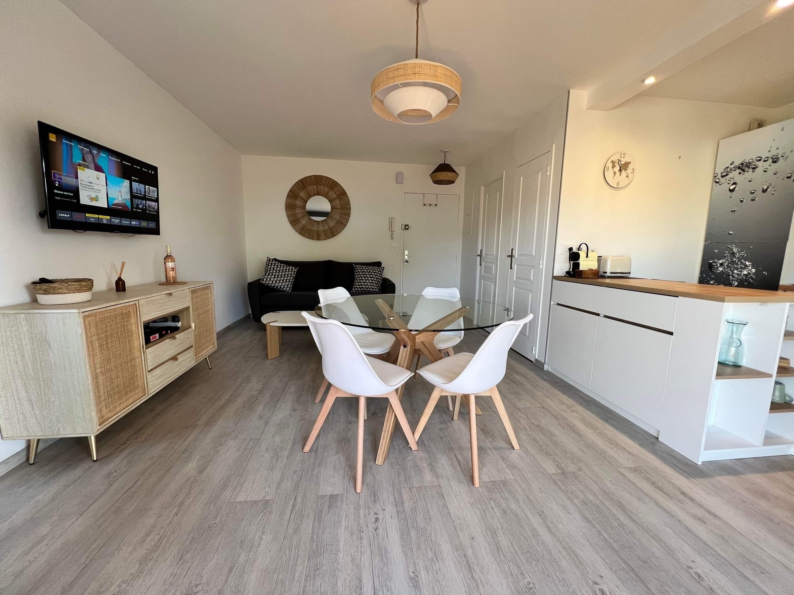 A modern living room with a glass dining table, white chairs, a black sofa, and a TV mounted on the wall.