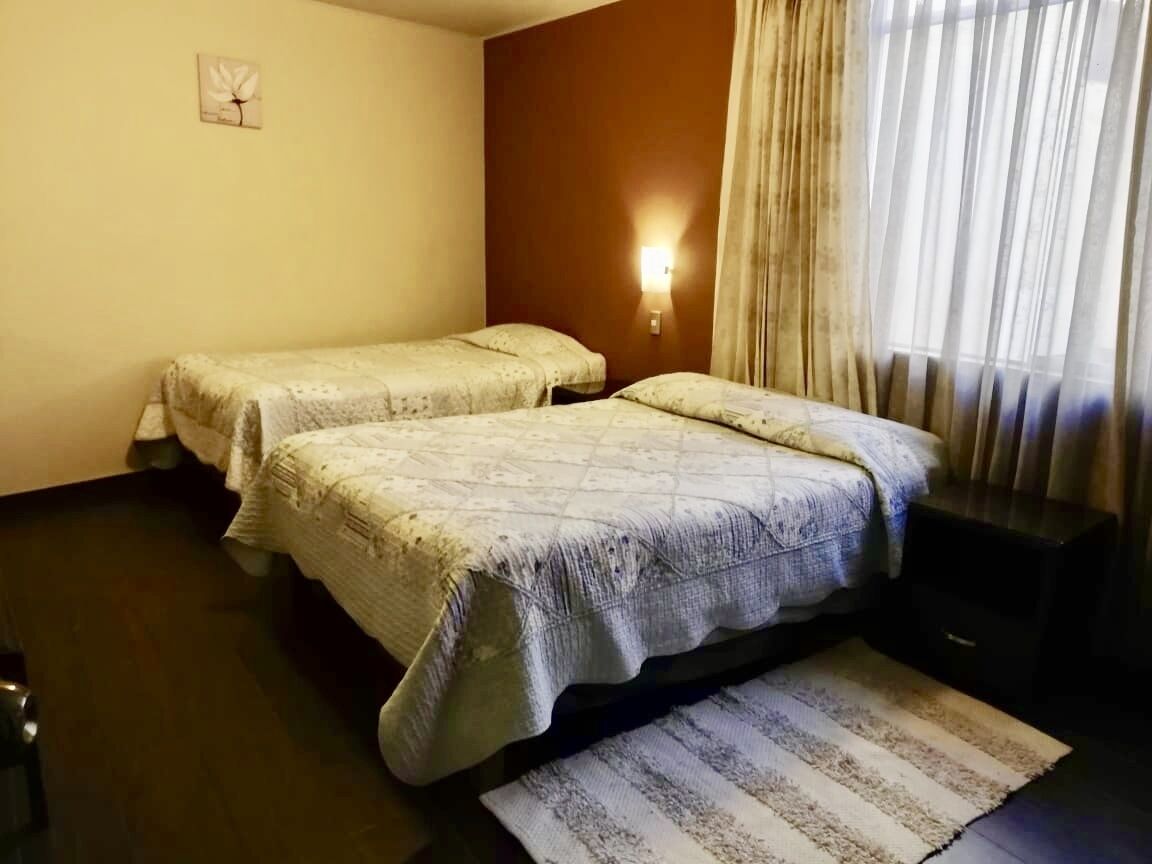 A hotel room with two beds, a small rug, a wall clock, and a window with curtains.