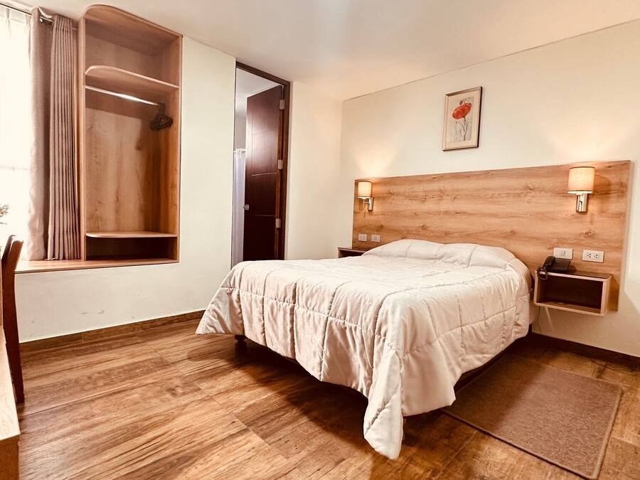 Habitación de hotel con piso de madera, cama con cubrecama blanco, mesita de noche con lámpara, armario de madera y ventana con cortinas.