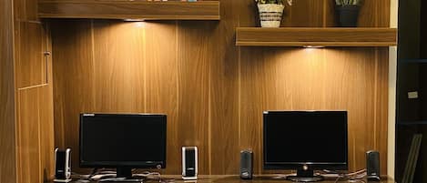A wooden-paneled room with two desks, each with a computer, keyboard, and mouse. There are speakers on either side of the desks, and a chair in front of each. A bookshelf with books and a potted plant is above the desks.